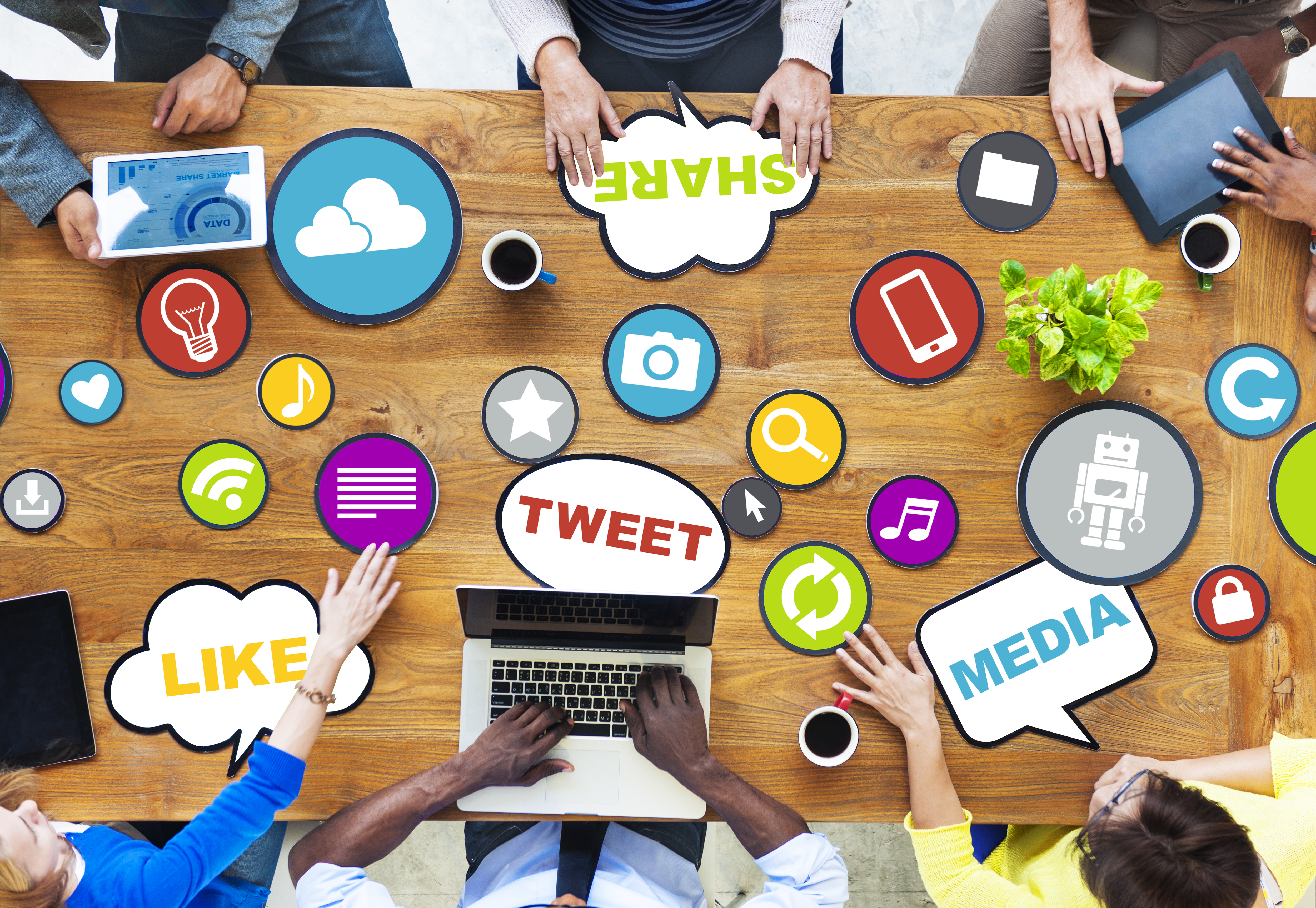 top view photo of six people sitting at a table with laptops and tablets with animated words around the table such as tweet, share, like and media